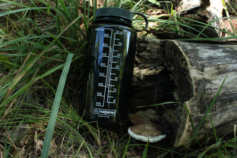 The forest-green Nalgene 32oz water bottle, with a white liquid measurement scale (in milliliters and in ounces) on the back of it, next to a fallen log and mushroom, in a grassy shaded area. The text on the bottle reads: "For your everyday adventures. Nalgene. Made in USA."