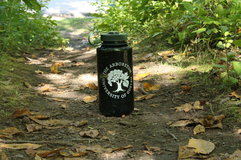 The forest-green Nalgene 32oz water bottle, with a white Arboretum logo on it, on a trail path, admidst fallen leaves and plants.