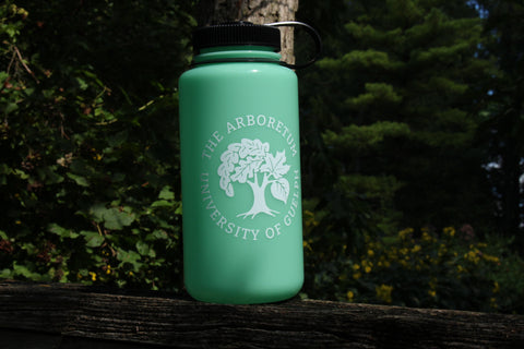The glow-green Nalgene 32oz water bottle, with a white Arboretum logo on it, sitting on top of a wooden fence in the sun, with trees in the background.