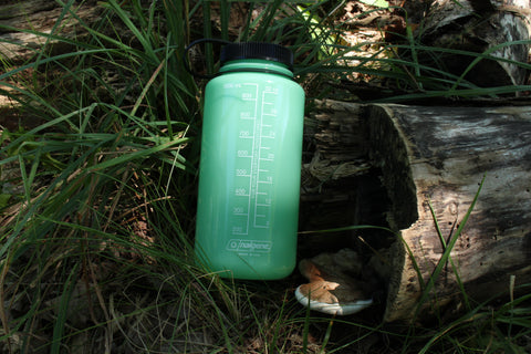 The glow-green Nalgene 32oz water bottle, with a white liquid measurement scale (in milliliters and in ounces) on the back of it, next to a fallen log and mushroom, in a grassy shaded area. The text on the bottle reads: "For your everyday adventures. Nalgene. Made in USA."