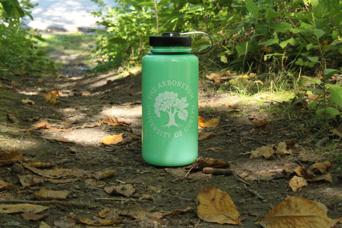 The glow-green Nalgene 32oz water bottle, with a white Arboretum logo on it, on a trail path, admidst fallen leaves and plants.