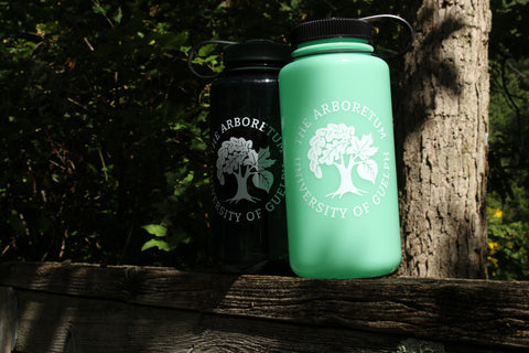 The forest-green and glow-green Nalgene 32oz water bottles, with white Arboretum logos on them, on top of a wooden fence, with trees in the background.