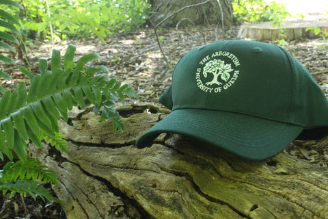A forest-green ball cap hat, with an embroidered Arboretum tree logo and text "The Arboretum, University of Guelph" on the front. The hat sits on a log with a fern growing next to it.
