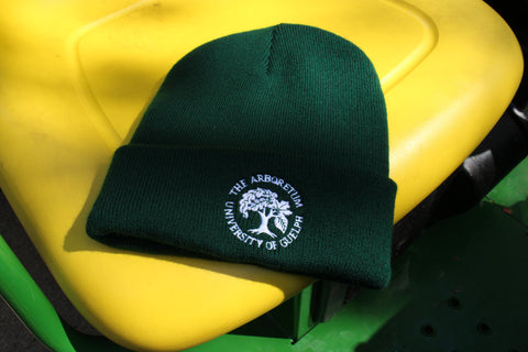 A forest-green beanie winter hat, with an embroidered Arboretum tree logo and text "The Arboretum, University of Guelph" on the front brim. The hat sits on a bright yellow Gator vehicle seat.