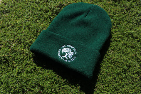 A forest-green beanie winter hat, with an embroidered Arboretum tree logo and text "The Arboretum, University of Guelph" on the front brim. The hat sits in a sunny patch of Creeping Thyme. 