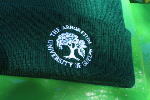 A close-up of the embroidered Arboretum tree logo and text "The Arboretum, University of Guelph" on the front brim of the forest-green beanie hat. The hat sits on a shiny green background.