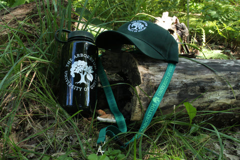 A forest-green Arboretum ball cap hat sitting on a fallen log, with a green Arboretum lanyard hung around the log. Next to the log, a forest-green Arboretum Nalgene 32oz water bottle sits in the grass.