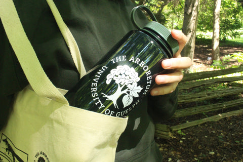  A person's hand pulls the forest-green Nalgene 32oz water bottle, with a white Arboretum logo on it, out of an Arboretum tote bag. The background is a forested area with an old wooden fence.