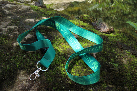 Green lanyard with 'University of Guelph Arboretum' text on one side, and a centimeters ruler on the other side, on a mossy rock, with frogs behind it.